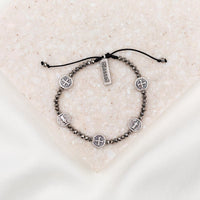 A silver metallic crystal blessing bracelet with adjustable cording, featuring silver-tone Benedictine medals and a 'blessed' charm tag, displayed on a textured background