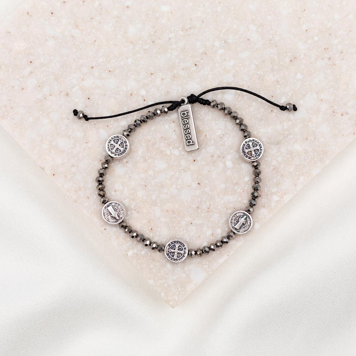 A silver metallic crystal blessing bracelet with adjustable cording, featuring silver-tone Benedictine medals and a 'blessed' charm tag, displayed on a textured background