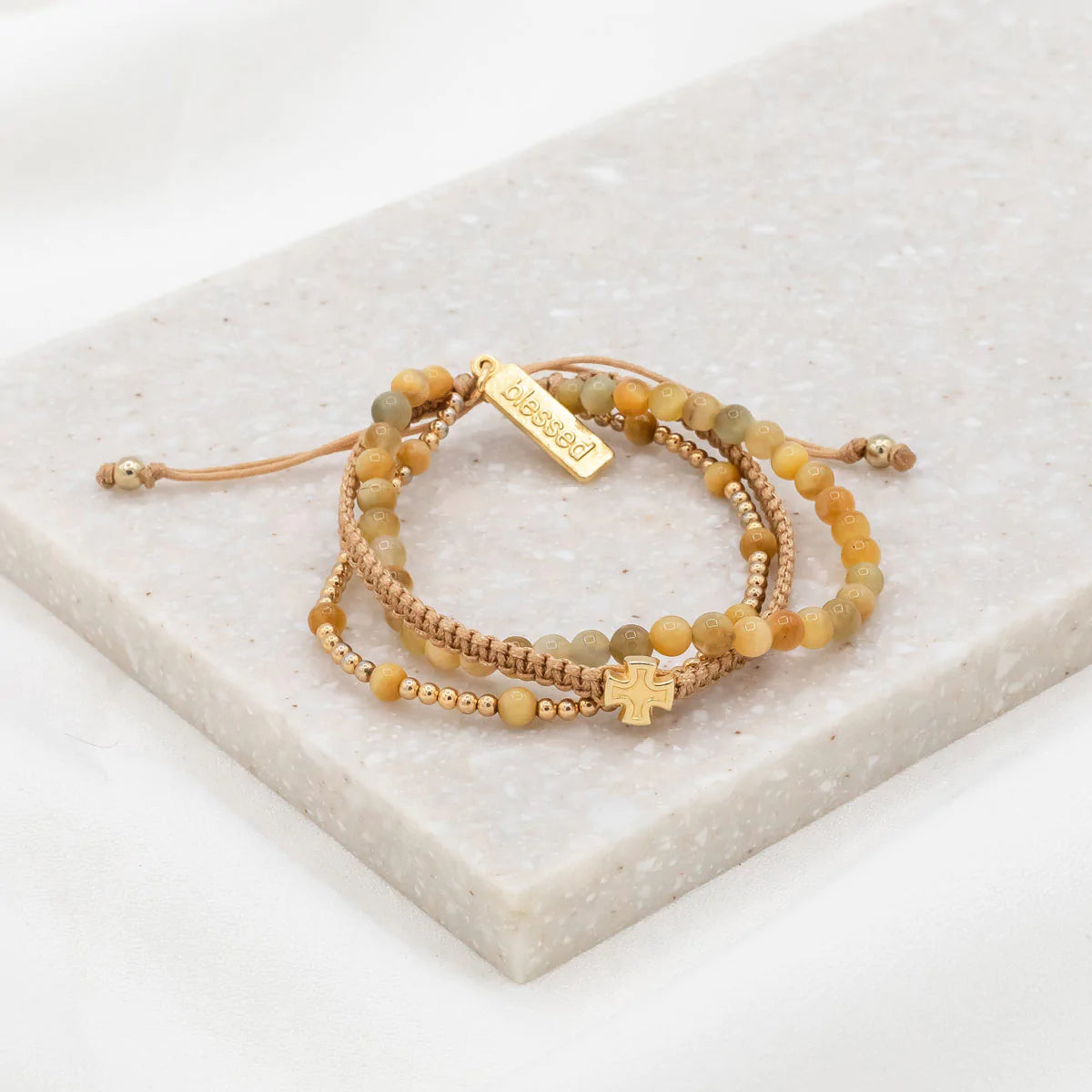 A set of three handwoven bracelets made with Tiger's Eye gemstones, gold-tone beads, and a cross medal, displayed on a marble surface.