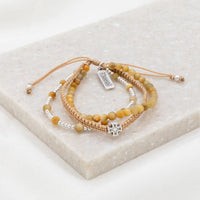 A set of three handwoven bracelets made with Tiger's Eye gemstones, silver-tone beads, and a cross medal, displayed on a marble surface.