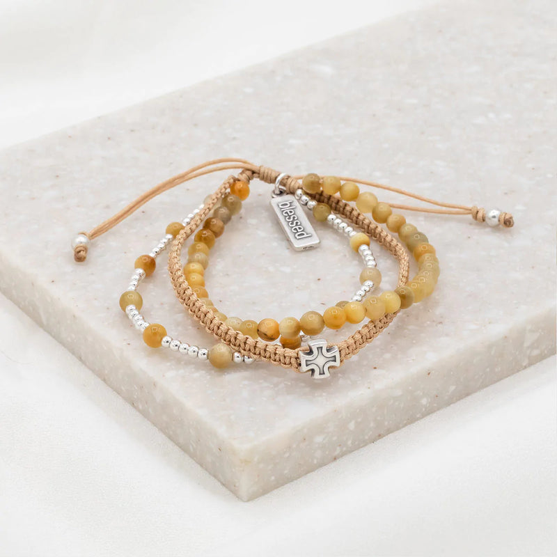 A set of three handwoven bracelets made with Tiger's Eye gemstones, silver-tone beads, and a cross medal, displayed on a marble surface.