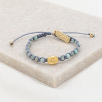 A Miraculous Medal bracelet with blue beads and a gold-tone medal, displayed on a beige surface.