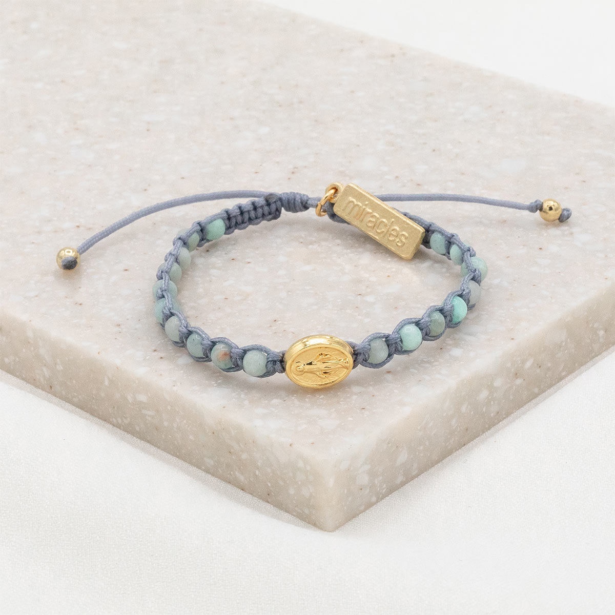 A Miraculous Medal bracelet with blue beads and a gold-tone medal, displayed on a beige surface.