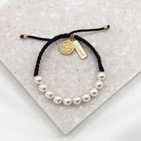 A hand wearing a gold-tone bracelet with a Benedictine medal and a 'blessed' charm tag, featuring white beads and a gold-colored cord.