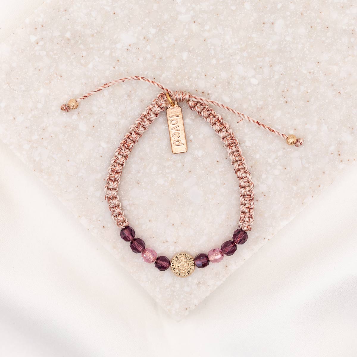 An adjustable bracelet with pink and purple crystals and a gold-tone St. Amos medal, displayed on a white surface.