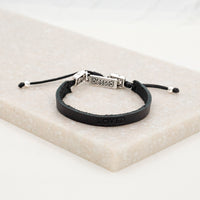 A black leather bracelet with metal clasps, stamped with the word 'LOVED', accompanied by two silver-tone Auspice Maria/Sacred Heart of Jesus Medals and a 'blessed' charm tag, displayed on a white marble surface.