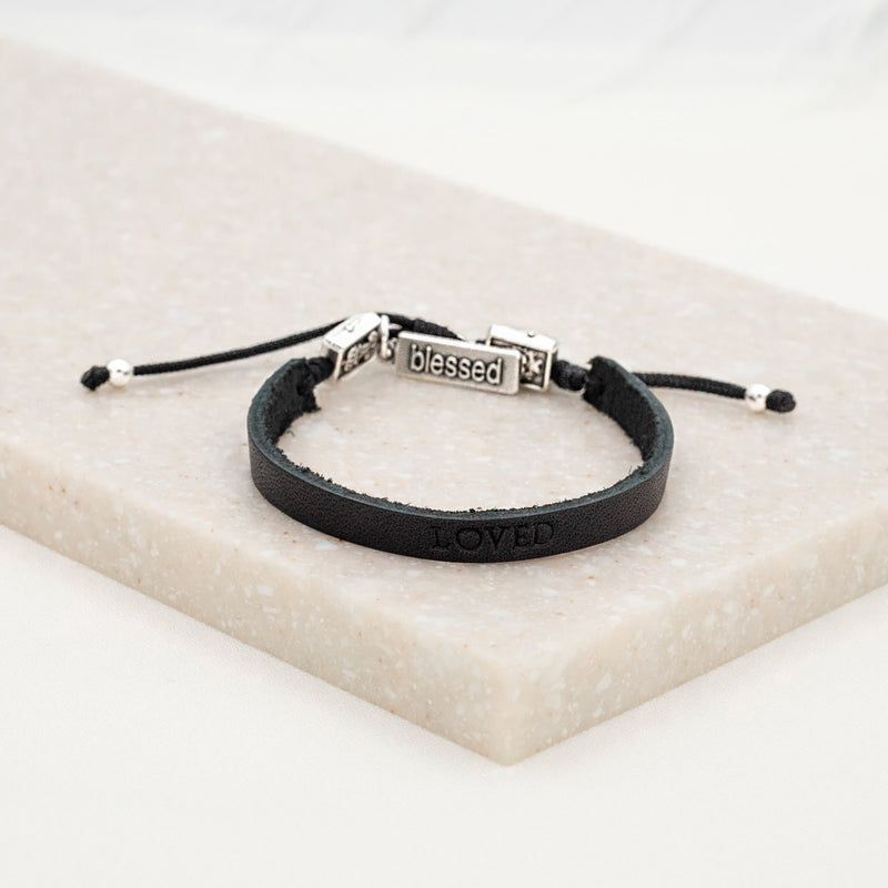A black leather bracelet with metal clasps, stamped with the word 'LOVED', accompanied by two silver-tone Auspice Maria/Sacred Heart of Jesus Medals and a 'blessed' charm tag, displayed on a white marble surface.