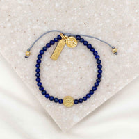 A bracelet with dark blue beads and gold-tone charms including a medal of St. Francis and a 'loved' tag, displayed on a white surface.