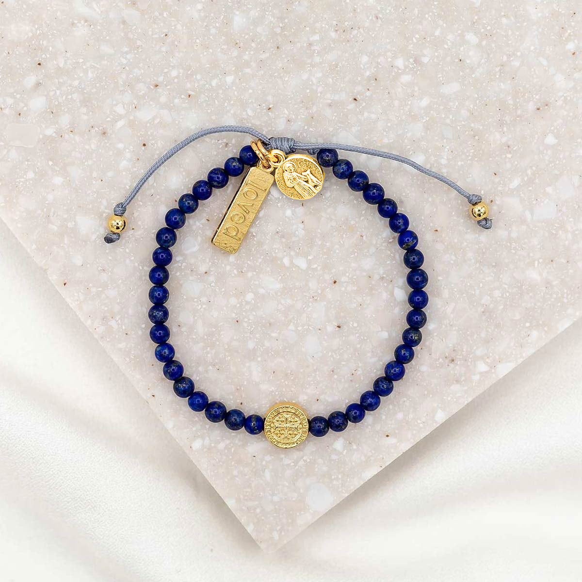 A bracelet with dark blue beads and gold-tone charms including a medal of St. Francis and a 'loved' tag, displayed on a white surface.