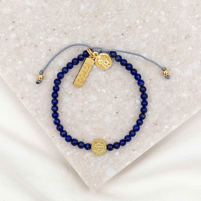 A bracelet with dark blue beads and gold-tone charms including a medal of St. Francis and a 'loved' tag, displayed on a white surface.