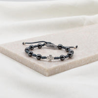 A handwoven bracelet with silver-tone beads, including a cross and a 'blessed' tag, displayed on a white marble surface.