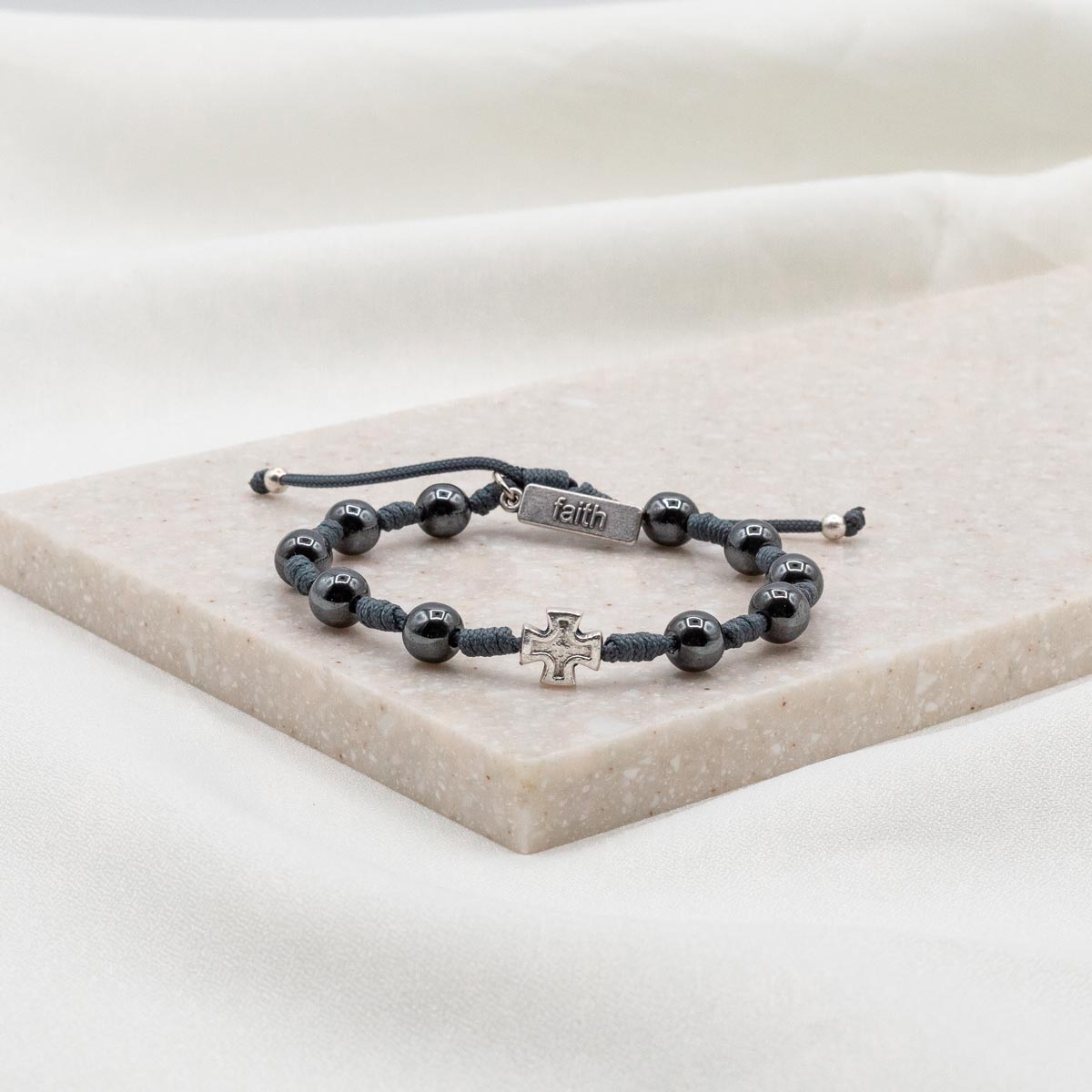 A handwoven bracelet with silver-tone beads, including a cross and a 'blessed' tag, displayed on a white marble surface.