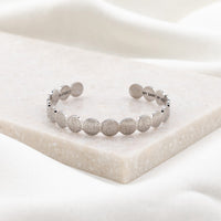 A silver-plated stainless steel cuff bracelet with embossed text, displayed on a white marble surface.