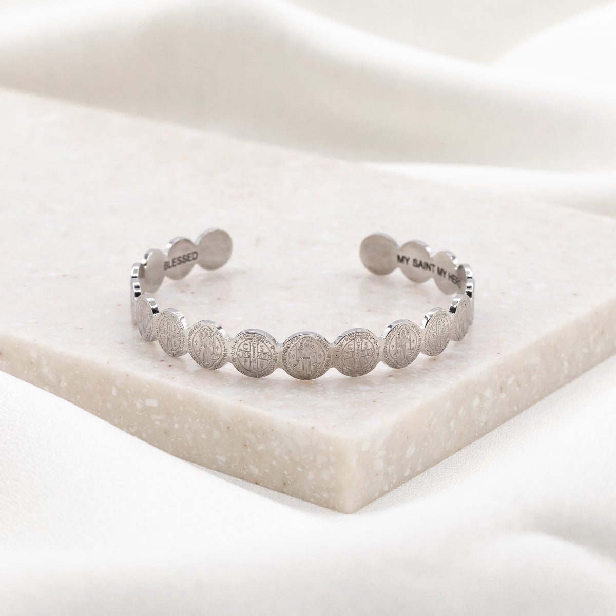A silver-plated stainless steel cuff bracelet with embossed text, displayed on a white marble surface.