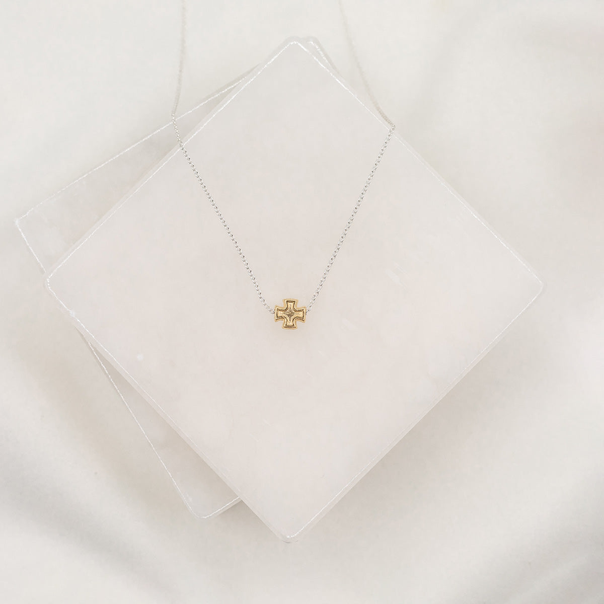 A petite cross necklace with a silver dipped chain, displayed on a white cushion.