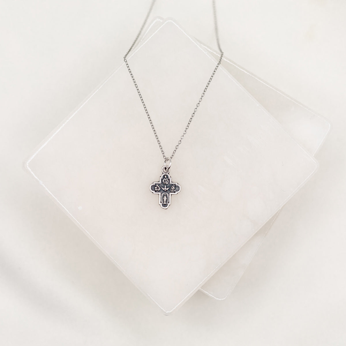 A silver-tone dainty cross necklace on a white display stand.
