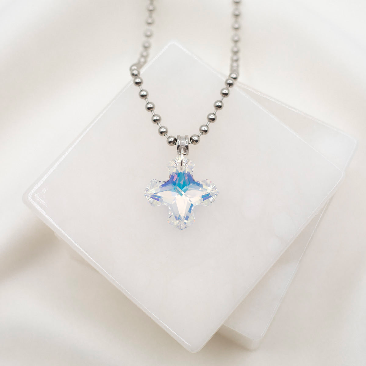 A silver-colored cross necklace with a large, clear, square-shaped crystal pendant, displayed against a white background.