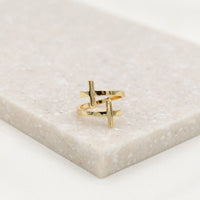 A gold-dipped cross ring placed on a beige surface, with an adjustable design.