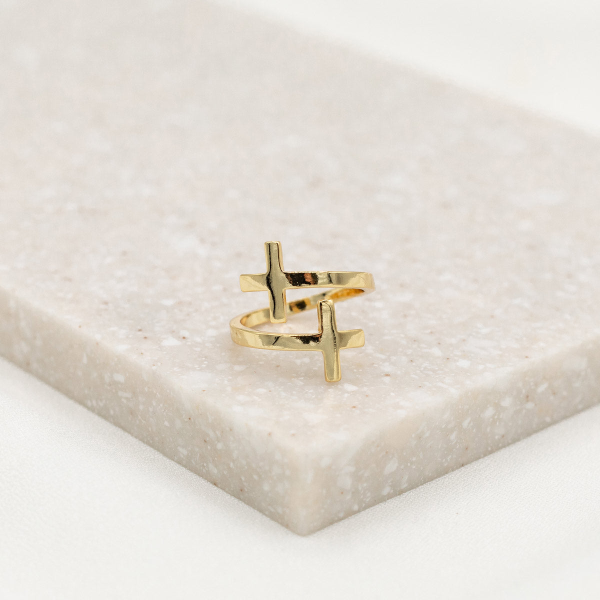A gold-dipped cross ring placed on a beige surface, with an adjustable design.