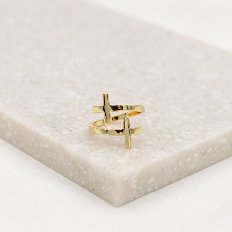A gold-dipped cross ring placed on a beige surface, with an adjustable design.