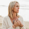 Writer Sara Schulting Kranz wearing 7 Stones of Forgiveness gemstone dainty bracelet with her hands over her heart, looking off to distance, foggy day at beach