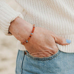 Women's hand wearing Be Kind Humankind 7 Stones of Forgiveness carnelian and aquamarine gemstone beaded dainty bracelet, woman's hand in denim jeans pocket with blue nail polish and cream woven sweater