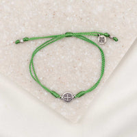 A green bracelet with a silver-tone Benedictine medal and a logo charm, displayed on a white surface.