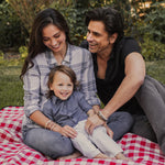 John Stamos and Caitlin Stamos with their son wearing their My Saint My Hero Share the Love St. Amos Collection Bracelets, outside on grass on checked red and white picnic blanket
