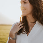 Young brunette woman facing right with the sunrise behind her, wearing white shirt, a glory saints and heroes silver charm beaded bracelet and a blessings and grace benedictine necklace with gemstones and crystals