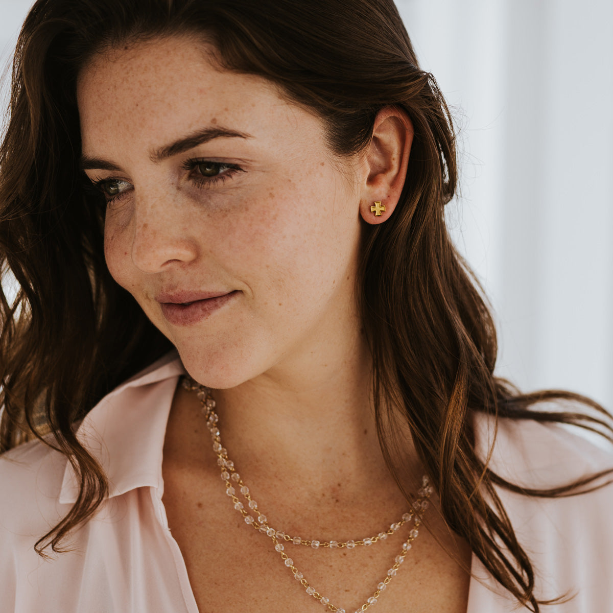 Young woman wearing gold tone faith petite cross earrings and a my saint my hero rosary bead style layer of light necklace