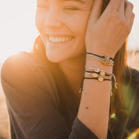 young woman smiling, sun over her shoulder, wearing stack of petite Mary Say Yes string bracelets