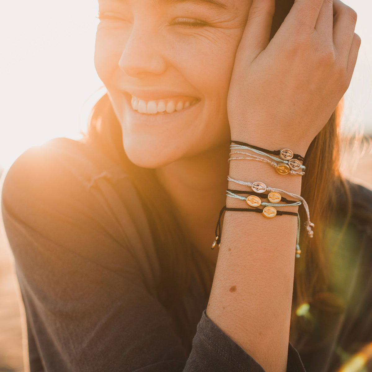 young woman smiling, sun over her shoulder, wearing stack of petite Mary Say Yes string bracelets